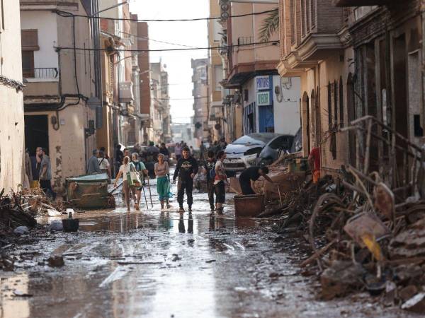 Unas personas caminan por una calle cubierta de lodo y rodeada de coches amontonados tras las intensas lluvias por la fuerte DANA, en Valencia.