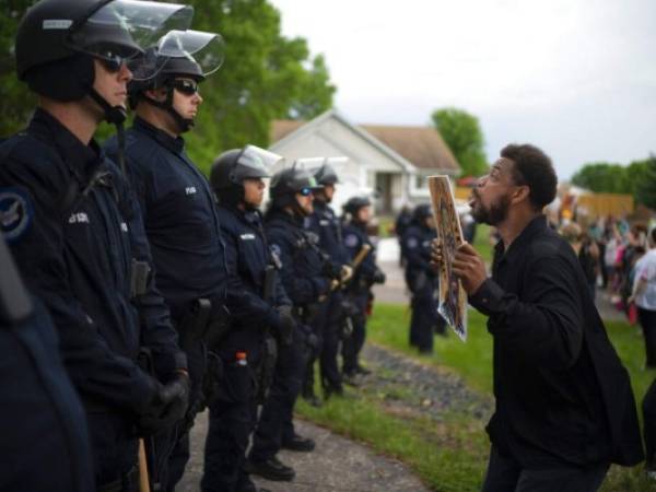 Un manifestante sostiene una pancarta mientras salta en repetidas ocasiones para que los policías que se encuentran al fondo puedan verlo en la casa del agente Derek Chauvin, quien fue despedido de la policía de Minneapolis, a las afueras de Oakdale, Minnesota. Foto: AP.